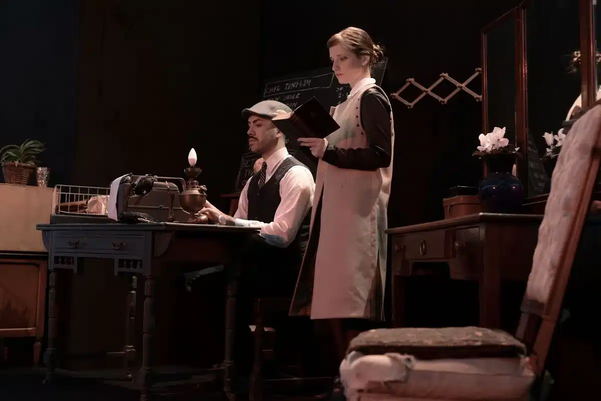 Une jeune femme en robe d'époque écrivant à un bureau, avec une expression déterminée et réfléchie.